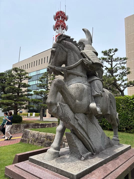 福井城址に立つ結城秀康の騎馬像。石造りの馬にまたがる甲冑姿の武者で、背後に県庁舎と松の木、赤白の鉄塔が見える